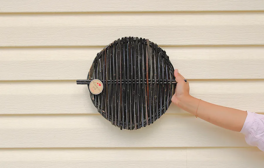 A round tray woven from dark willow rods, held up against a cream farmhouse wall